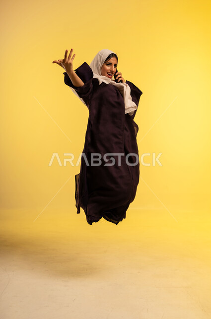 الدردشة مع الاصدقاء عبر الجوال، استخدام جهاز تقني حديث ومتطور، بورتريه لامرأة عربية خليجية عمانية ترتدي العباءة والحجاب  تمسك بيدها الهاتف المحمول تقوم بإجراء مكالمة هاتفية، شابة سعودية تقفز عاليا بإيماءات الفرح لسماع اخبار جيدة، الجسم بالطول الكامل، خلفية صفراء
