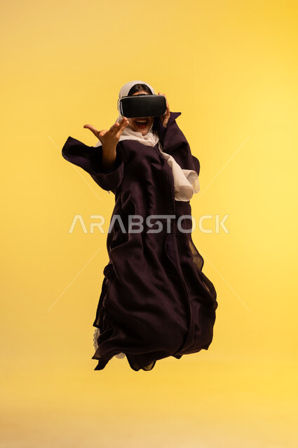 المتعة والتسلية بالمشاهدة عبر نظارة الواقع الافتراضي, بورتريه لامرأة عربية خليجية عمانية ترتدي العباءة والحجاب تضع نظارات ثلاثية الأبعاد، مشاهدة الافلام والمسلسلات، شابة سعودية تقفز عاليا بتعابير الاندماج ومحاكاة العالم الخيالي، الجسم بالطول الكامل، خلفية صفراء