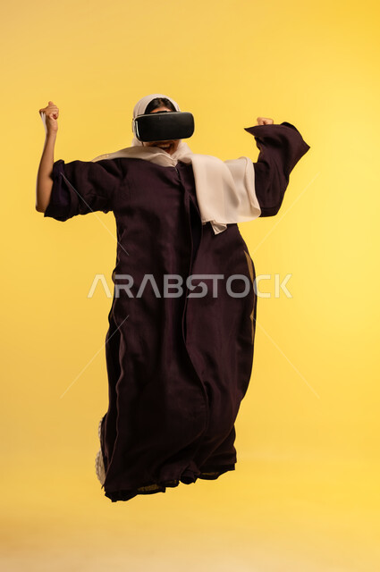 Watching movies and series, fun and entertainment by watching through virtual reality glasses, portrait of an Arab Gulf Omani woman wearing an abaya and hijab, wearing 3D glasses, a young Saudi woman jumping high with expressions of integration and simulating the imaginary world, full-length body, yellow background