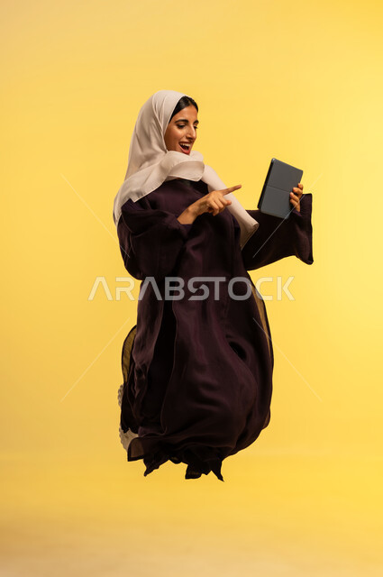 Integrating technology into work, watching movies and series via tablet, using a modern and advanced technical device, portrait of an Arab Gulf Omani woman wearing an abaya and hijab jumping high using a tablet, a young Saudi woman completing the required tasks remotely, full body, yellow background