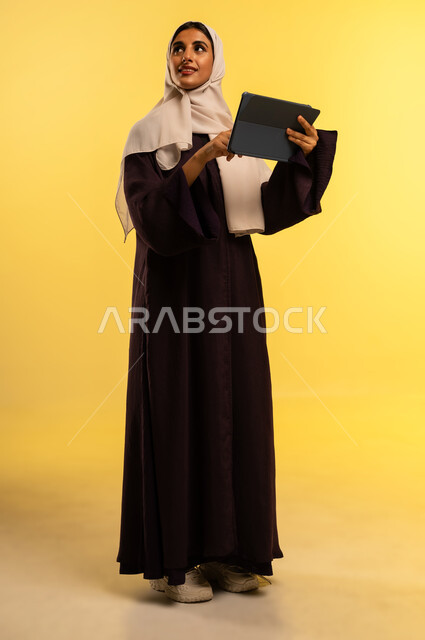 استخدام جهاز تقني حديث ومتطور، بورتريه لامرأة عربية خليجية عمانية ترتدي العباءة والحجاب تستخدم الجهاز اللوحي، شابة سعودية تقوم بإنجاز المهام المطلوبة عن بعد، مشاهدة الافلام والمسلسلات عبر التابلت، دمج التكنولوجيا بالعمل، الجسم بالطول الكامل، خلفية صفراء