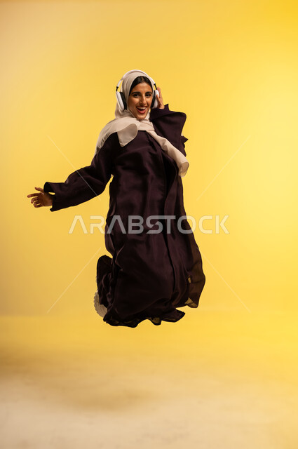 الاندماج مع الأنغام الموسيقية، الانصات والاستماع للأغاني باستخدام الهيدفون، بورتريه لامرأة عربية خليجية عمانية ترتدي العباءة والحجاب تستمع إلى الموسيقى من خلال سماعة الرأس، شابة سعودية تقفز عاليا تنظر إلى الكاميرا بإيماءات السرور، الجسم بالطول الكامل، خلفية صفراء
