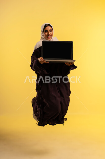 Integrating technology into work, completing required tasks remotely, using a modern and advanced technical device, portrait of a smiling Omani Gulf Arab woman wearing an abaya and hijab jumping high using a laptop, a young Saudi woman showing a blank black screen via laptop, full body, yellow background