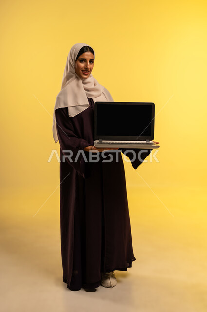 إنجاز المهام المطلوبة عن بعد، استخدام جهاز تقني حديث ومتطور، بورتريه لامرأة عربية خليجية عمانية مبتسمة ترتدي العباءة والحجاب تستخدم الحاسوب المحمول، شابة سعودية تعرض شاشة سوداء فارغة عبر اللابتوب، دمج التكنولوجيا بالعمل، الجسم بالطول الكامل، خلفية صفراء