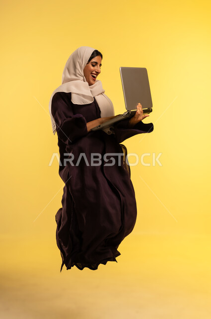 دمج التكنولوجيا بالعمل، إنجاز المهام المطلوبة عبر اللابتوب، استخدام جهاز تقني حديث ومتطور، بورتريه لامرأة عربية خليجية عمانية مبتسمة ترتدي العباءة والحجاب تقفز عاليا تستخدم الحاسوب المحمول، شابة سعودية تقوم بادارة الأعمال عن بعد، الجسم بالطول الكامل، خلفية صفراء