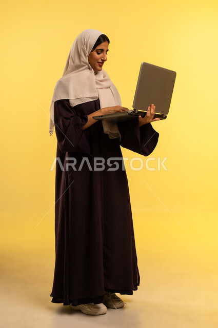 Female profession and job, using modern and advanced technical device, portrait of an Arab Gulf Omani woman wearing abaya and hijab using laptop, young Saudi woman managing business remotely, integrating technology into work, completing required tasks via laptop, full body, yellow background