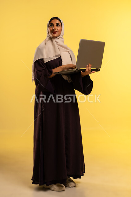 استخدام جهاز تقني حديث ومتطور، بورتريه لامرأة عربية خليجية عمانية ترتدي العباءة والحجاب تستخدم الحاسوب المحمول، شابة سعودية تقوم بادارة الأعمال عن بعد، دمج التكنولوجيا بالعمل، إنجاز المهام المطلوبة عبر اللابتوب، مهنة ووظيفة نسائية، الجسم بالطول الكامل، خلفية صفراء
