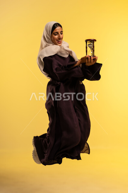 تحديد مواعيد العمل وتنظيم الجدول الزمني، مفهوم إدارة الأعمال والأوقات، شابة سعودية تقفز عاليا بتعابير السرور، بورتريه لامرأة عربية خليجية عمانية ترتدي الحجاب والعباءة تحمل الساعة الرملية الزجاجية (البنكام)، أهميه مراقبة الوقت, الجسم بالطول الكامل، خلفية صفراء