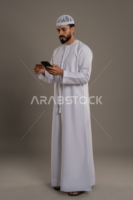 إمكانية الدفع الالكتروني باستخدام البطاقة، النظر إلى الهاتف المحمول بإيماءات التركيز، الشراء والدفع من خلال برامج الجوال، بورتريه لرجل عربي خليجي اماراتي يرتدي الكندورة والقبعة البيضاء يمسك بيده الهاتف المحمول وبطاقة الإئتمان البنكية، خلفية رمادية