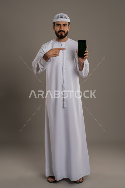عرض تطبيقات وخدمات الهاتف المحمول، الاشارة إلى الجوال بشاشة سوداء فارغة، استخدام التكنولوجيا المتطورة للتسويق الرقمي الالكتروني، بورتريه لرجل عربي خليجي اماراتي يرتدي الكندور وينظر إلى الكاميرا، صورة الجسم بالطول الكامل، خلفية رمادية
