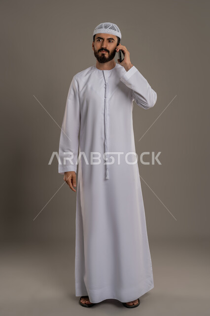 Elegance and interest in appearance, using mobile phone to communicate and connect with individuals, searching for work online, portrait of an Arab Gulf Emirati man wearing a kandura and making a phone call via mobile, full body image, gray background