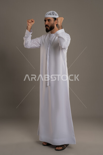 الحماس والسعادة العارمة، الفرح  بتحقيق الأهداف والأحلام، النجاح والفوز بشيء ما منتظر، بورتريه لرجل عربي خليجي اماراتي مبتسم يرتدي الكندورة والطاقية يرفع قبضتيه في الهواء بإيماءات السرور، الجسم بالطول الكامل، خلفية رمادي