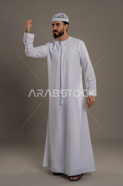 الفرح  بتحقيق الأهداف والأحلام، الحماس والسعادة العارمة، النجاح والفوز بشيء ما منتظر، بورتريه لرجل عربي خليجي اماراتي مبتسم يرتدي الكندورة والطاقية يرفع قبضته في الهواء بإيماءات السرور، الجسم بالطول الكامل، خلفية رمادي