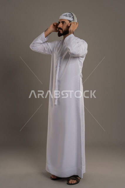 مفهوم الإصغاء والاستمتاع بإستخدام الهيدفون، النظر إلى مكان ما بايماءات الطرب والتركيز،  قضاء أوقات ممتعة بالإستماع للمواهب الغنائية، بورتريه لرجل عربي خليجي اماراتي يرتدي الكندورة ويضع سماعة الرأس، الجسم بالطول الكامل، خلفية رمادي
