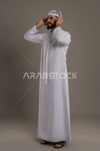 النظر إلى مكان ما بايماءات الطرب والتركيز، مفهوم الإصغاء والاستمتاع بإستخدام الهيدفون، قضاء أوقات ممتعة بالإستماع للمواهب الغنائية، بورتريه لرجل عربي خليجي اماراتي يرتدي الكندورة ويضع سماعة الرأس، الجسم بالطول الكامل، خلفية رمادي