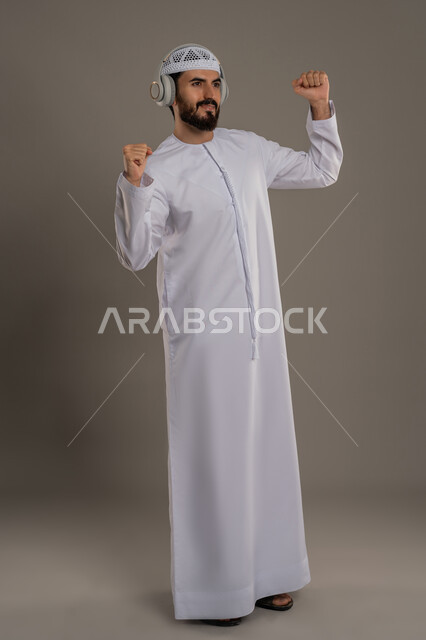 النظر إلى مكان ما بايماءات الطرب والتركيز، مفهوم الإصغاء والاستمتاع بإستخدام الهيدفون، قضاء أوقات ممتعة بالإستماع للمواهب الغنائية، بورتريه لرجل عربي خليجي اماراتي يرتدي الكندورة ويضع سماعة الرأس، الجسم بالطول الكامل، خلفية رمادي