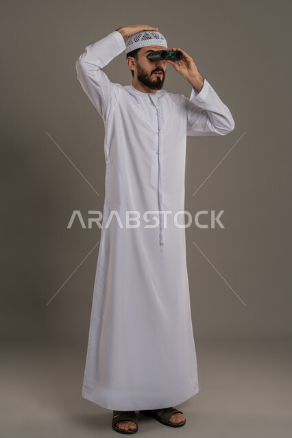 التخطيط ووضع الأهداف المستقبلية، استخدام المنظار ثنائي العينين لمشاهدة الأشياء البعيدة، توضيح الرؤية وتقريبها، بورتريه لرجل عربي خليجي اماراتي يرتدي الكندورة والطاقية يقف ويضع تلسكوب العدسات المقربة على عينيه يضع يده على راسه، الجسم بالطول الكامل، خلفية رمادي