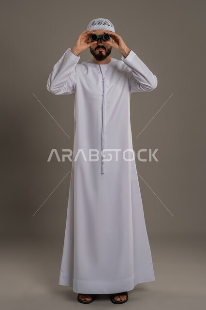 استخدام المنظار ثنائي العينين لمشاهدة الأشياء البعيدة، توضيح الرؤية وتقريبها، التخطيط ووضع الأهداف المستقبلية بورتريه لرجل عربي خليجي اماراتي يرتدي الكندورة والطاقية يقف ويضع تلسكوب العدسات المقربة على عينيه، الجسم بالطول الكامل، خلفية رمادي