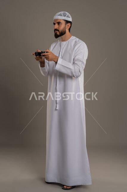 استخدام الأجهزة التكنولوجية والتقنيات الحديثة، تعابير الاستمتاع باللعب بالألعاب الالكترونية الترفيهية، بورتريه لرجل عربي خليجي اماراتي يرتدي الكندورة والغترة يجلس يقف ويمسك بيده عصا التحكم بإيماءات التركيز والاندماج، الجسم بالطول الكامل، خلفية رمادي