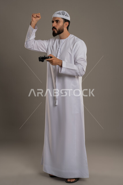 تعابير الاستمتاع باللعب بالألعاب الالكترونية الترفيهية، استخدام الأجهزة التكنولوجية والتقنيات الحديثة، بورتريه لرجل عربي خليجي اماراتي يرتدي الكندورة والطاقية يقف ويمسك بيده عصا التحكم بإيماءات التركيز والاندماج، الجسم بالطول الكامل، خلفية رمادي