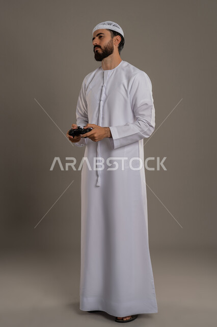 Using technological devices and modern techniques, expressions of enjoyment playing entertaining electronic games, portrait of an Arab Gulf Emirati man wearing a kandura and a ghutra, sitting, standing and holding a joystick in his hand with gestures of focus and integration, full-length body, gray background