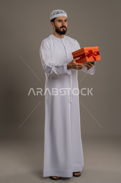 تقديم الهدايا بالاعياد والمناسبات الخاصة، مفهوم الشكر والامتنان، التعبير عن الحب والتقدير والاحترام، بورتريه لرجل عربي خليجي اماراتي يرتدي الكندورة والطاقية يحمل صندوق هدية وينظر إلى مكان ما بايماءات السرور، الجسم بالطول الكامل، خلفية رمادي
