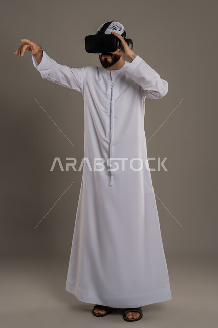 محاكاة العالم الخيالي, استخدام حاسة اللمس لملامسة عناصر هولوجرامية بإيماءات النقر, بورتريه لرجل عربي خليجي اماراتي يرتدي الكندورة والطاقية ويضع نظارة الواقع الافتراضي ثلاثية الأبعاد يشير إلى شيء ما بأصبع السبابة, صورة الطول الكامل للجسم, خلفية رمادي