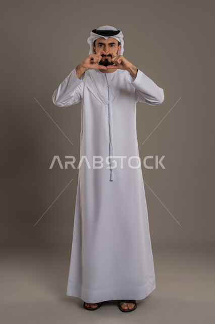 إظهار مشاعر الحب و الشغف، مفهوم المحبة و المودة, النظر إلى الكاميرا بإيماءات السعادة و السرور,  بورتريه لرجل عربي خليجي اماراتي يرتدي الكندورة التقليدية والغترة يشكل بيديه شكل قلب,  الجسم بالطول الكامل، خلفية رمادي