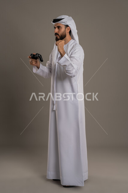 تعابير الاستمتاع باللعب بالألعاب الالكترونية الترفيهية، استخدام الأجهزة التكنولوجية والتقنيات الحديثة، بورتريه لرجل عربي خليجي اماراتي يرتدي الكندورة والغترة يقف ويمسك بيده عصا التحكم بإيماءات الفوز والحماس، الجسم بالطول الكامل، خلفية رمادي