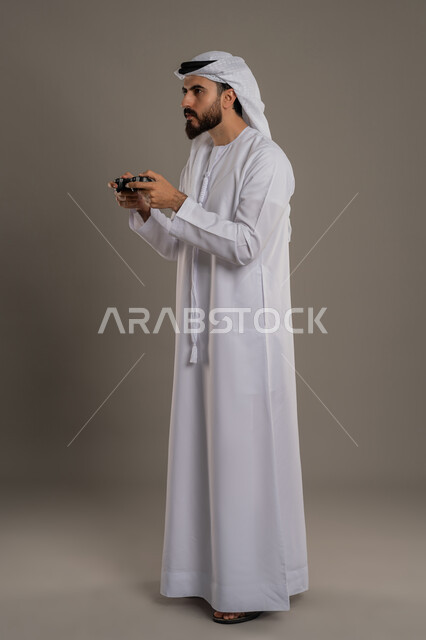 تعابير الاستمتاع باللعب بالألعاب الالكترونية الترفيهية، استخدام الأجهزة التكنولوجية والتقنيات الحديثة، بورتريه لرجل عربي خليجي اماراتي يرتدي الكندورة والغترة  يقف ويمسك بيده عصا التحكم بإيماءات التركيز والاندماج، الجسم بالطول الكامل، خلفية رمادي