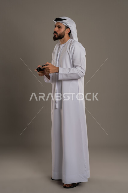 استخدام الأجهزة التكنولوجية والتقنيات الحديثة، تعابير الاستمتاع باللعب بالألعاب الالكترونية الترفيهية، بورتريه لرجل عربي خليجي اماراتي يرتدي الكندورة والغترة يقف ويمسك بيده عصا التحكم بإيماءات التركيز والاندماج، الجسم بالطول الكامل، خلفية رمادي