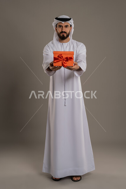 مفهوم الشكر والامتنان، تقديم الهدايا بالاعياد والمناسبات الخاصة، التعبير عن الحب والتقدير والاحترام، بورتريه لرجل عربي خليجي اماراتي يرتدي الكندورة والغترة يحمل صندوق هدية  وينظر إلى الكاميرا بايماءات السرور، الجسم بالطول الكامل، خلفية رمادي