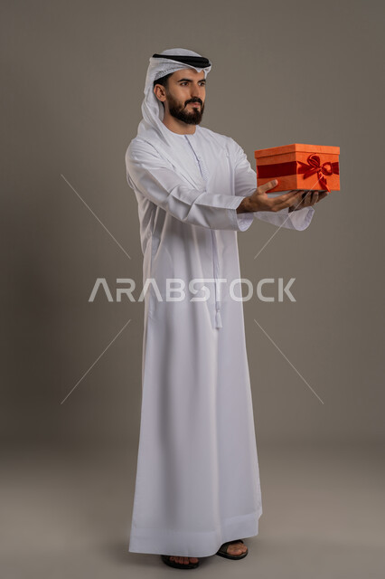 Concept of thanks and gratitude, giving gifts on holidays and special occasions, expressing love, appreciation and respect, portrait of an Arab Gulf Emirati man wearing a kandura and ghutra holding a gift box and looking at the camera with gestures of pleasure, full body, gray background