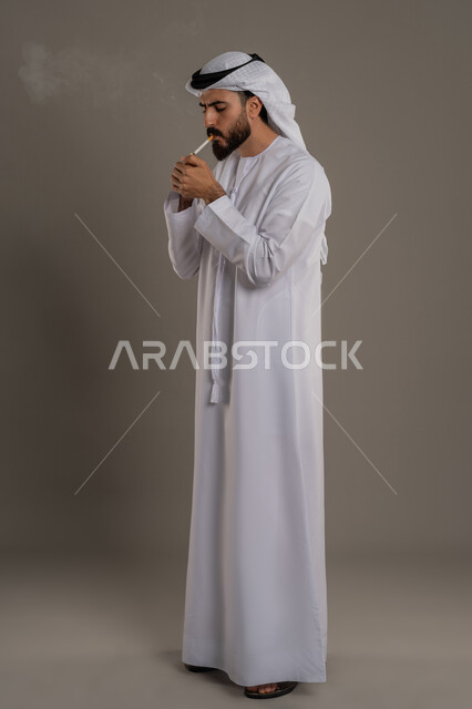 عادات سلوكية خاطئة، الادمان على التبغ والسجائر، تعاطي مواد ضارة وسامة، الإصابه بأمراض الجهاز التنفسي، بورتريه لرجل عربي اماراتي خليجي يرتدي الكندورة والغترة يقوم بسحب نفس عميق من السيجارة، الجسم بالطول الكامل، خلفية رمادي