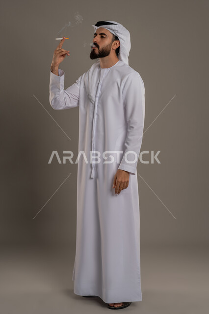الادمان على التبغ والسجائر، عادات سلوكية خاطئة، تعاطي مواد ضارة وسامة، الإصابه بأمراض الجهاز التنفسي، بورتريه لرجل عربي اماراتي خليجي يرتدي الكندورة والغترة يقوم بسحب نفس عميق من السيجارة، الجسم بالطول الكامل، خلفية رمادي