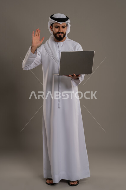 عقد إجتماع عمل عبر الإنترنت، تطور التكنولوجيا والأجهزة التقنية، استخدام الحاسوب المحمول لإنجاز المهام، بورتريه لرجل عربي اماراتي خليجي يرتدي الكندورة والغترة يقف ويحمل جهاز اللابتوب يرفع كف يديه بايماءات القاء التحية  والسلام، الجسم بالطول الكامل، خلفية رمادية