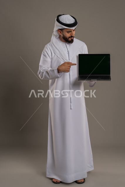 الاشارة باصبع السبابة إلى شاشة سوداء فارغة، تصفح مواقع التواصل الإجتماعي والعمل عن بعد، استخدام جهاز تقني حديث ومتطور لانجاز المشاريع، بورتريه لرجل عربي خليجي اماراتي يرتدي الكندورة والغترة البيضاء يحمل بيده الحاسوب المحمول، خلفية رمادية