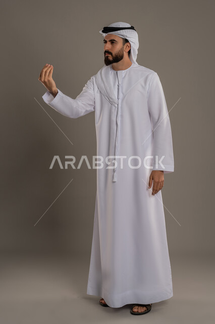 مفهوم الصبر والتريث و التأني, التهديد و التوعد لشيء ما، بورتريه لرجل عربي خليجي اماراتي يرتدي الثوب التقليدي والغترة ينظر الى الكاميرا يرفع يده نحو الأعلى بإيماءات طلب الإنتظار, إمهال بعض الوقت لفعل شيء، الطول الكامل للجسم, خلفية رمادي