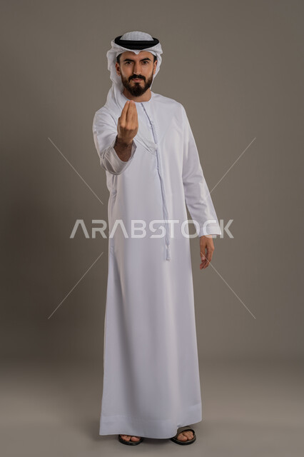 التهديد و التوعد لشيء ما، مفهوم الصبر والتريث و التأني,  بورتريه لرجل عربي خليجي اماراتي يرتدي الثوب التقليدي والغترة ينظر الى الكاميرا يرفع يده نحو الأعلى بإيماءات طلب الإنتظار, إمهال بعض الوقت لفعل شيء، الطول الكامل للجسم, خلفية رمادي
