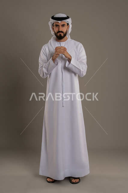 مفهوم الوسامة والثقة بالنفس، الوقوف والنظر إلى الكاميرا بإيماءات السرور والبشاشة، بورتريه لرجل عربي خليجي اماراتي مبتسم يرتدي الكندورة والغتره يقف ويشبك يديه ببعضهم، الرجولة العربية الأصيلة، الاهتمام بالمظهر الخارجي، الجسم بالطول الكامل، خلفية رمادي