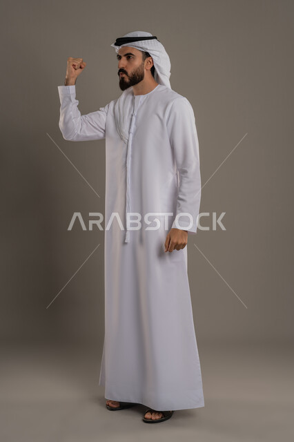 الفرح بتحقيق الأهداف والأحلام، الحماس والسعادة العارمة، النجاح والفوز بشيء ما منتظر، بورتريه لرجل عربي خليجي اماراتي يرتدي الكندورة والغترة يرفع قبضته في الهواء بإيماءات السرور، الجسم بالطول الكامل، خلفية رمادي