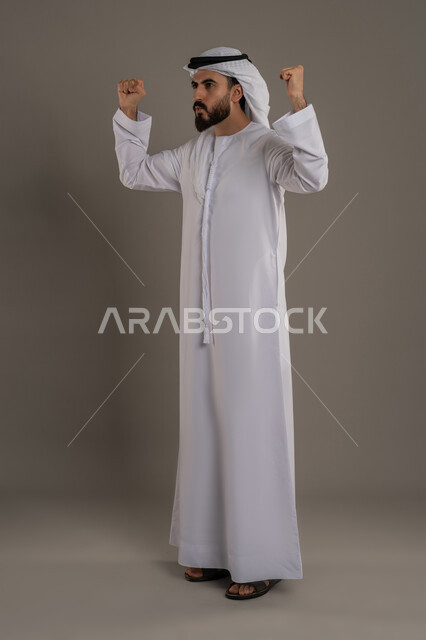النجاح والفوز بشيء ما منتظر، الفرح بتحقيق الأهداف والأحلام، الحماس والسعادة العارمة، بورتريه لرجل عربي خليجي اماراتي يرتدي الكندورة والغترة يرفع قبضته في الهواء بإيماءات السرور، الجسم بالطول الكامل، خلفية رمادي
