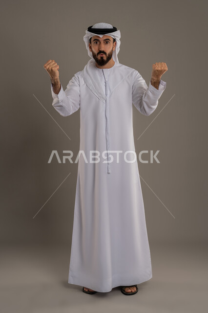 الحماس والسعادة العارمة، الفرح الهستيري بتحقيق الأهداف والأحلام، النجاح والفوز بشيء ما منتظر، بورتريه لرجل عربي خليجي اماراتي مبتسم يرتدي الكندورة والغترة يرفع قبضتيه في الهواء بإيماءات السرور، الجسم بالطول الكامل، خلفية رمادي