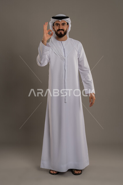 الامتياز والعلامة الكاملة، التعبير عن الإعجاب والتقدير، رفع اليد بعلامات الجودة والقبول، الشعور بالرضا والاستحسان، بورتريه لرجل عربي خليجي اماراتي يرتدي الكندورة والغترة ينظر إلى الكاميرا بإيماءات السعادة والسرور، الجسم بالطول الكامل، خلفية رمادي
