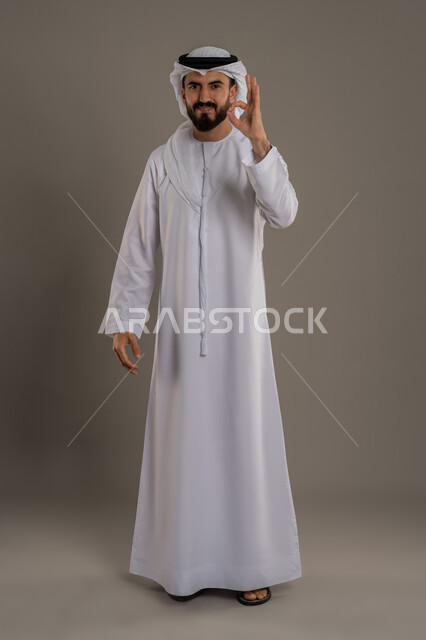 التعبير عن الإعجاب والتقدير، رفع اليد بعلامات الجودة والقبول، الامتياز والعلامة الكاملة، الشعور بالرضا والاستحسان، بورتريه لرجل عربي خليجي اماراتي يرتدي الكندورة والغترة ينظر إلى الكاميرا بإيماءات السعادة والسرور، الجسم بالطول الكامل، خلفية رمادي