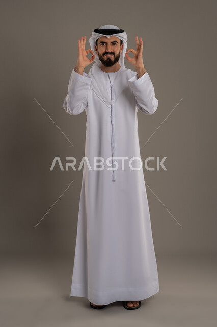 رفع اليد بعلامات الجودة والقبول، التعبير عن الإعجاب والتقدير، الامتياز والعلامة الكاملة، الشعور بالرضا والاستحسان، بورتريه لرجل عربي خليجي اماراتي يرتدي الكندورة والغترة ينظر إلى الكاميرا بإيماءات السعادة والسرور، الجسم بالطول الكامل، خلفية رمادي