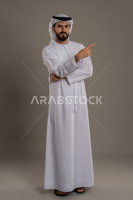 Looking at the camera with confident gestures, hand gestures indicating this place (here), locating a specific thing, portrait of an Arab Gulf Emirati man wearing a kandura and ghutra pointing with his index finger, interest in outward appearance, full body, white background