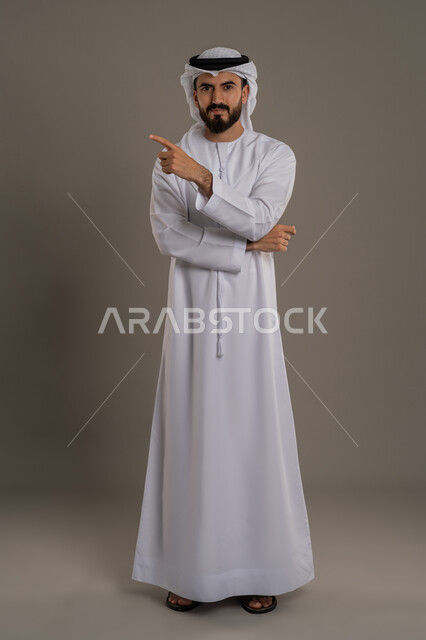 إشارات وحركات يدين تدل على هذا المكان (هنا)، النظر الى الكاميرا بايماءات الثقة بالنفس، تحديد موقع شيء معين، بورتريه لرجل عربي خليجي اماراتي يرتدي الكندورة والغترة يشير باصبع السبابة، الاهتمام بالمظهر الخارجي، الجسم بالطول الكامل، خلفية بيضاء