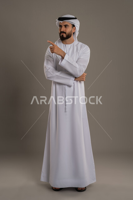 تحديد موقع شيء معين، إشارات وحركات يدين تدل على هذا المكان (هنا)، النظر الى مكان ما بايماءات الثقة بالنفس، بورتريه لرجل عربي خليجي اماراتي يرتدي الكندورة والغترة يشير باصبع السبابة، الاهتمام بالمظهر الخارجي، الجسم بالطول الكامل، خلفية بيضاء