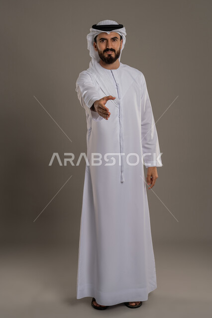 مد كف اليد نحو الأمام للمصافحة، إلقاء التحية والسلام، الأناقة والوسامة والرجولة، مفهوم التقدير والاحترام، بورتريه لرجل عربي خليجي اماراتي يرتدي الكندورة والغترة ينظر إلى الكاميرا بإيماءات السعادة والسرور، خلفية رمادي